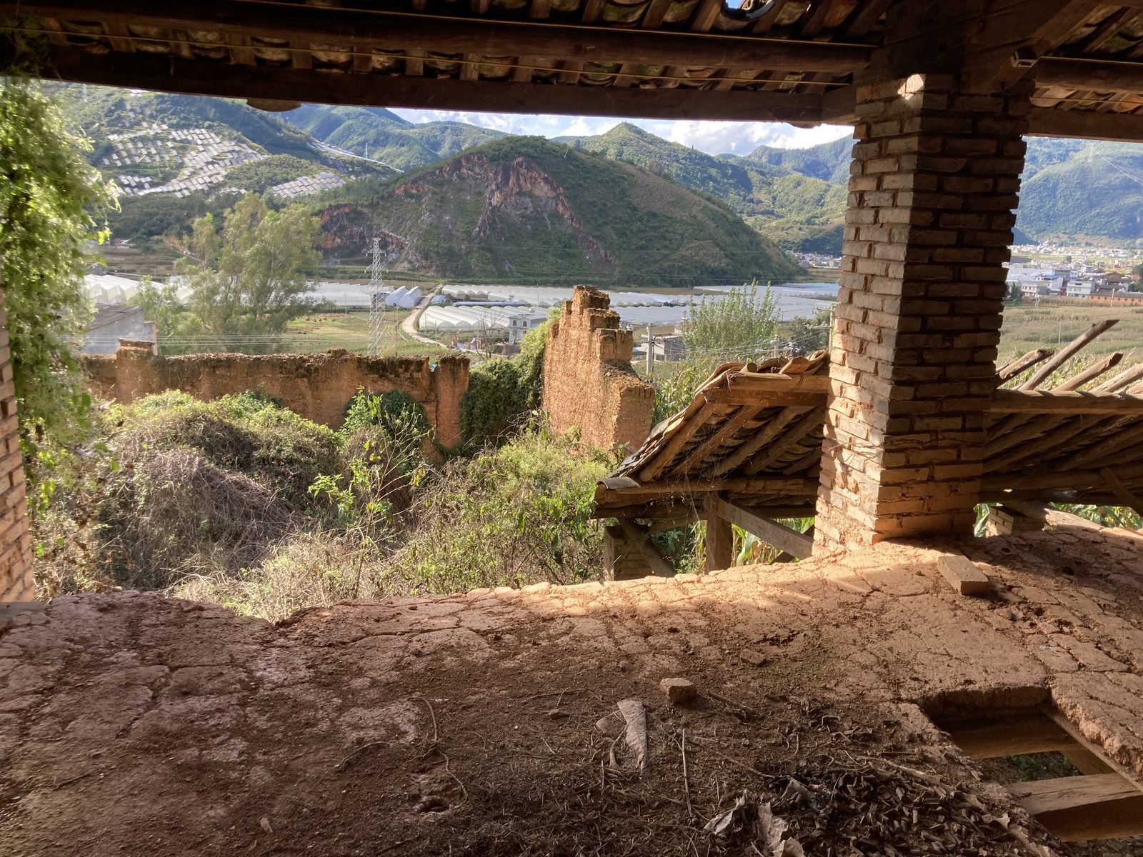 View from inside a ruined earth-walled building over a valley of greenhouses and terraced hills.